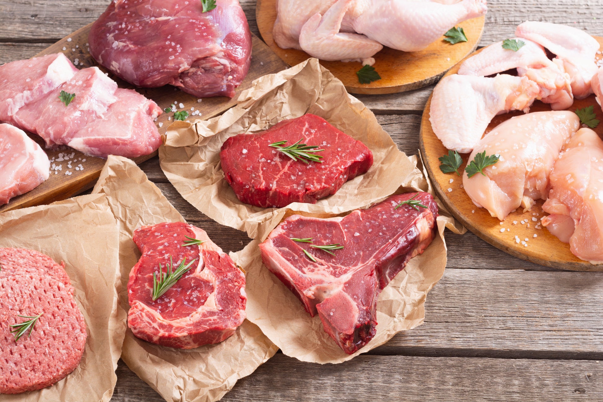 A variety of fresh meats including beef, pork, and chicken, some cut into steaks and others whole, displayed on a wooden board with parchment paper underneath.